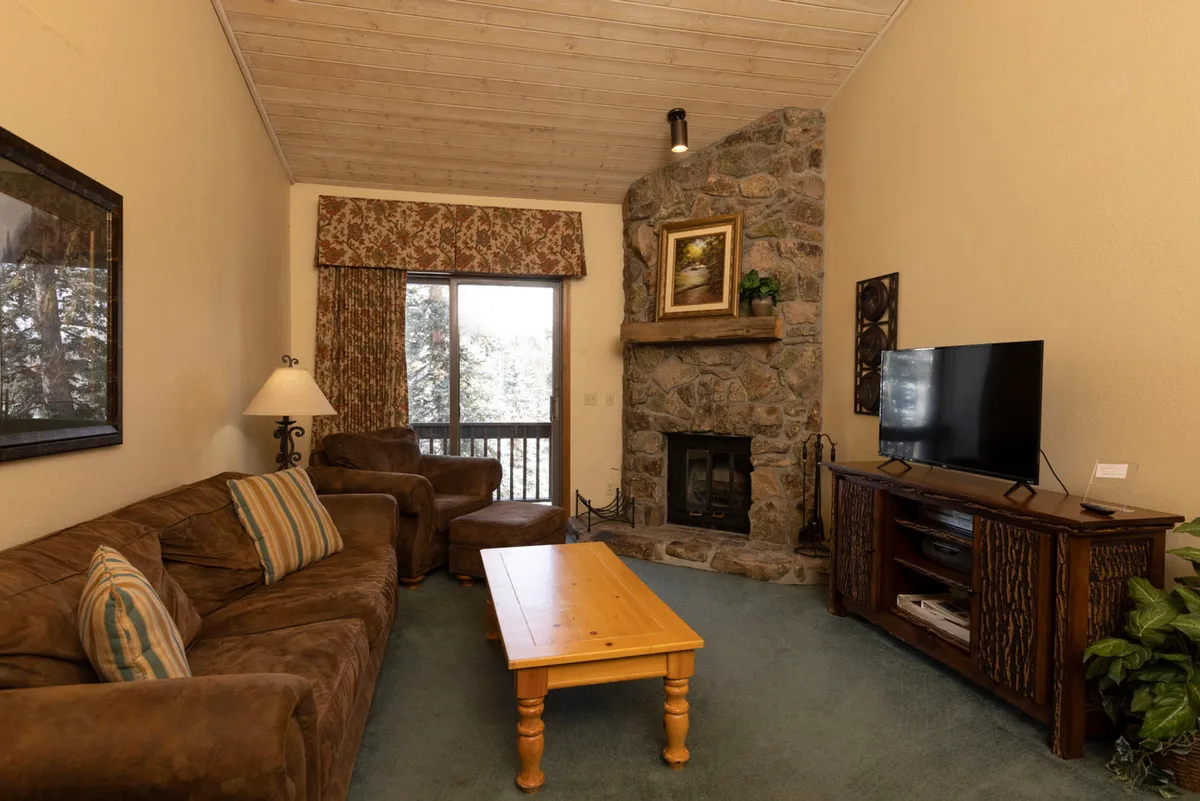 A warm and inviting living room featuring a rustic stone fireplace with a striking framed painting above, a plush brown suede sofa and armchair adorned with striped cushions, a central wooden coffee table, and a flat screen TV on a wooden stand—all set against soft lighting that creates a cozy, mountain retreat atmosphere.