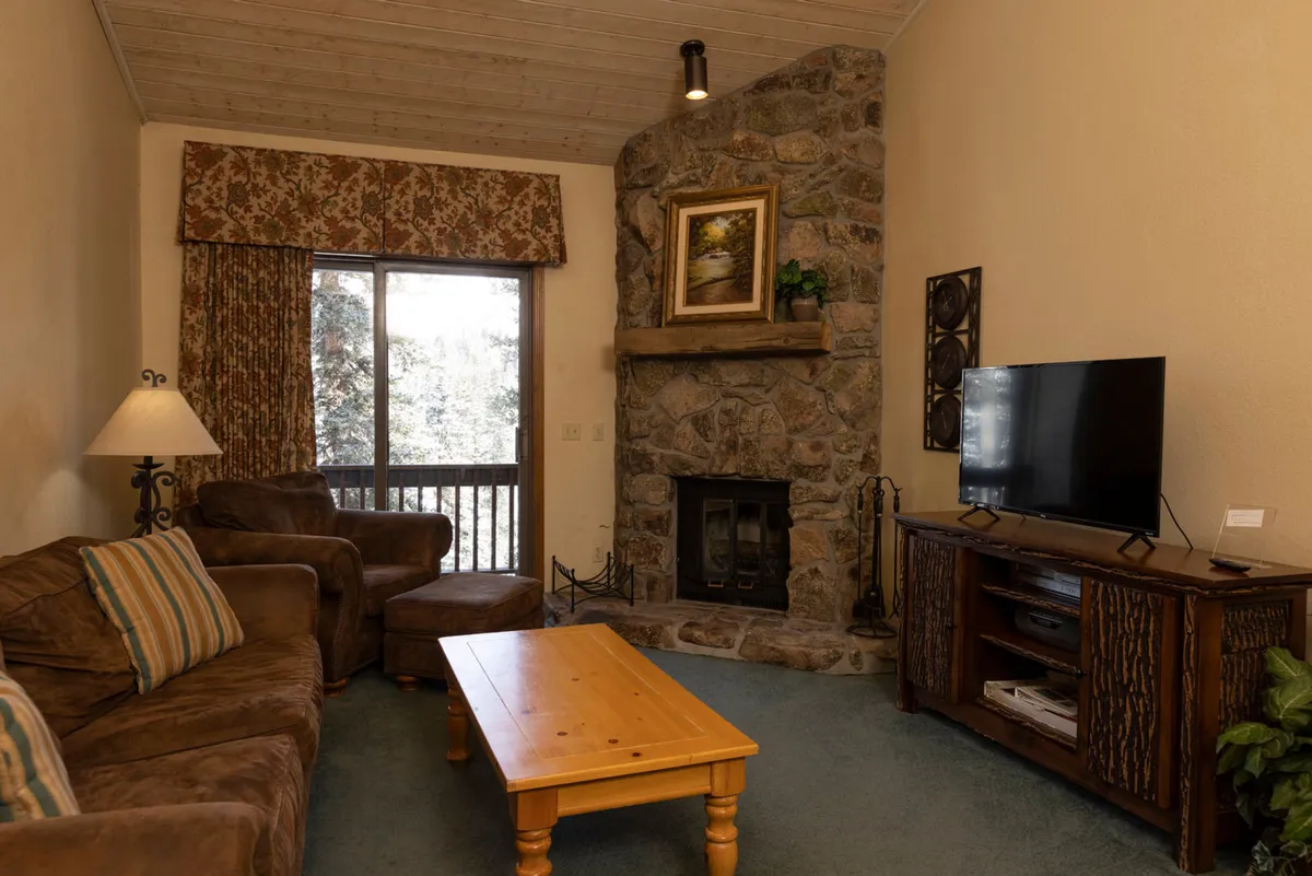 A cozy living room with a stone fireplace as the central feature, flanked by a wooden mantle and framed painting. The space includes a brown upholstered couch with striped cushions, an armchair, a rustic wooden coffee table, and a flat screen TV on a wooden stand. Large glass doors offer views outside, suggesting a connection to the outdoors.