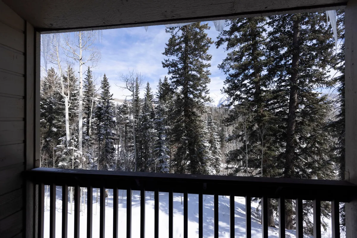 A wooden balcony overlooks a stunning view of snow-covered pine and aspen trees beneath a crisp blue sky. The scene is tranquil, inviting guests to enjoy serene winter mornings or unwind after a day on the mountain.