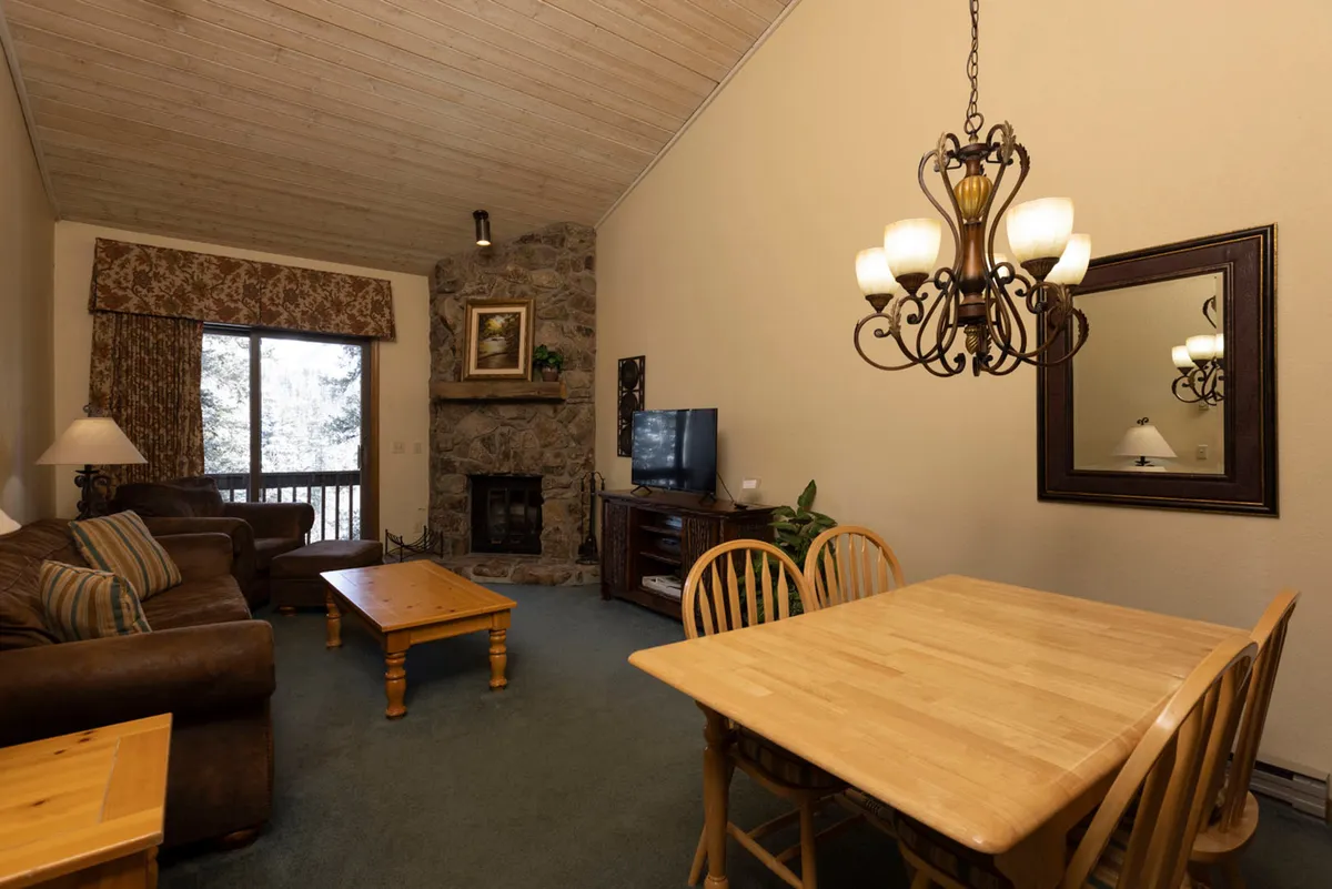 A spacious, open-concept living and dining area featuring a stone fireplace with a mantle and artwork, a wooden dining table with chairs, comfortable leather sofas, and a large window revealing snowy trees outside. The room is bathed in natural light, emphasizing the seamless flow between living and dining spaces.