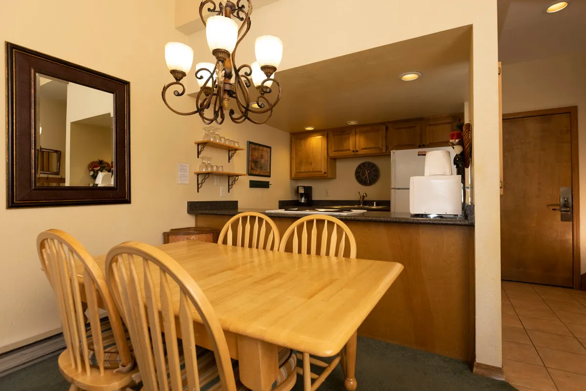 A warmly lit dining area with a wooden table and four chairs under an ornate chandelier. The kitchen counter in the background features a coffee maker and microwave, while a large mirror on the wall adds depth. The setting exudes comfort and is ideal for family meals or casual gatherings.