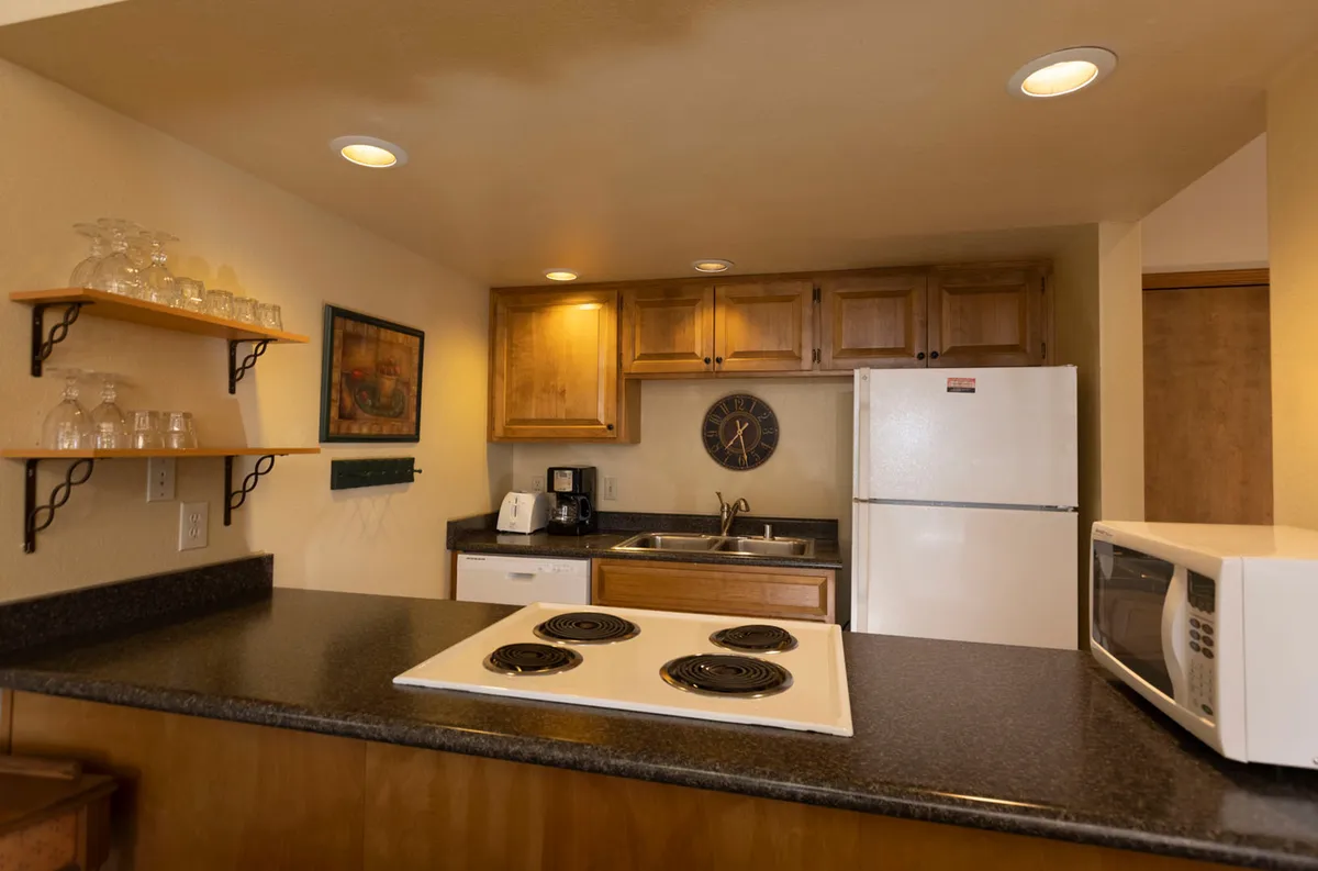A cozy kitchen featuring an electric stovetop on the counter, wooden cabinetry with overhead lighting, a white refrigerator, and open shelving stocked with glassware. The layout provides ample counter space, and the lighting creates a warm, inviting atmosphere ideal for meal preparation or casual dining.
