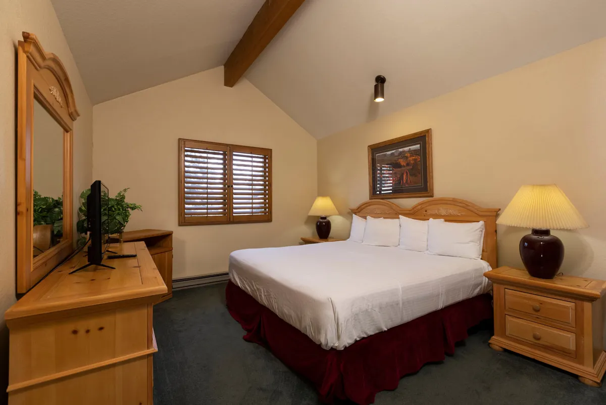 A cozy bedroom featuring a king-sized bed dressed in crisp white linens and a burgundy bed skirt. The room is accented by warm wooden furniture, including a dresser with a mirror and TV, matching nightstands with lamps, and a window with wooden shutters that lets in natural light.