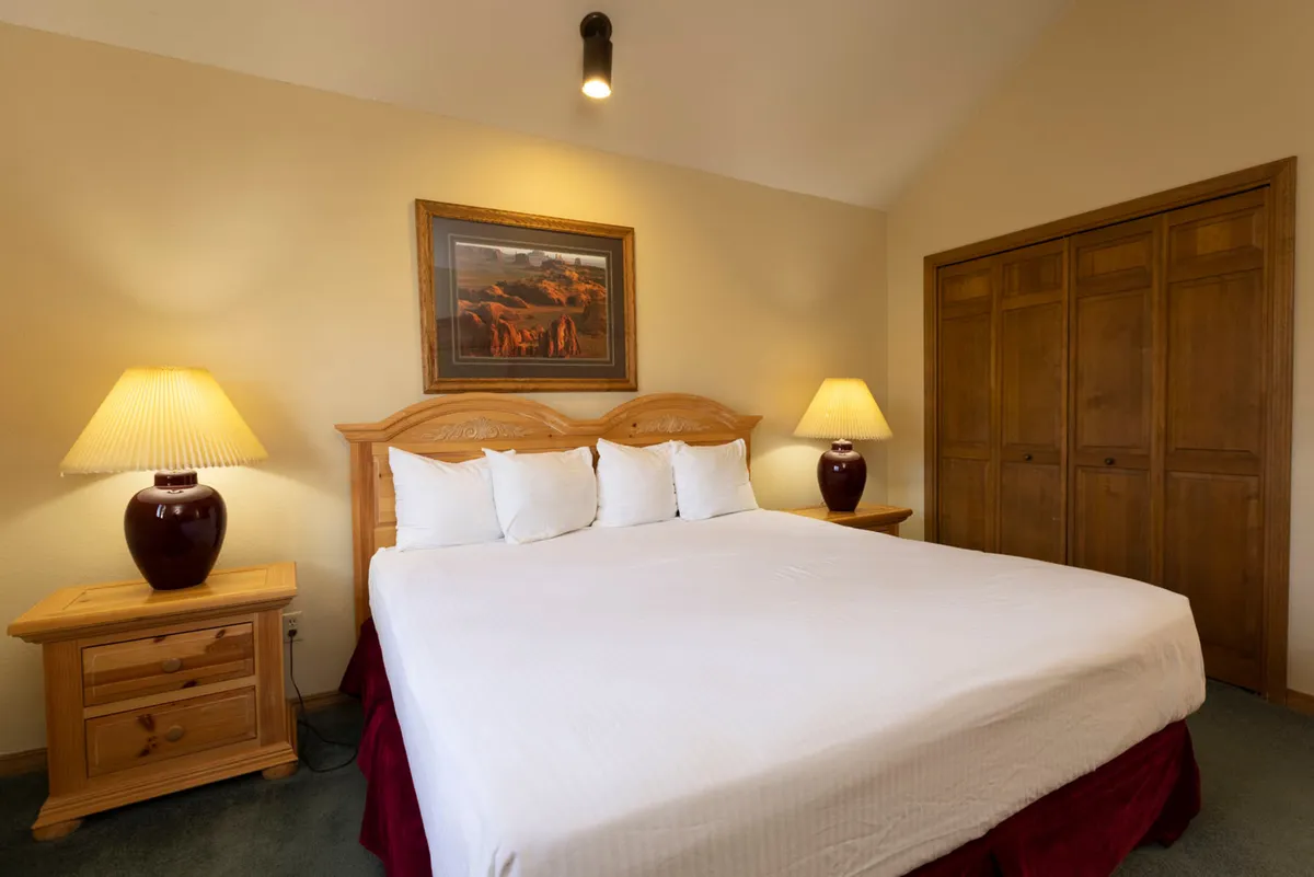 A warmly lit bedroom with a king-sized bed, crisp linens, matching wooden nightstands with lamps, a large wooden closet with paneled doors, and a serene landscape painting above the bed. The overall mood is restful and comfortable, ideal for unwinding after alpine adventures.