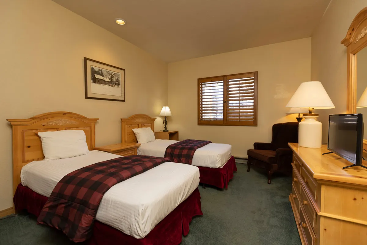 A charming twin bedroom with two beds adorned in plaid throws, a wooden dresser with a mirror and lamp, an armchair by the window, and wooden shutters framing the window. The overall ambiance is warm and inviting, perfect for family flexibility or accommodating guests.