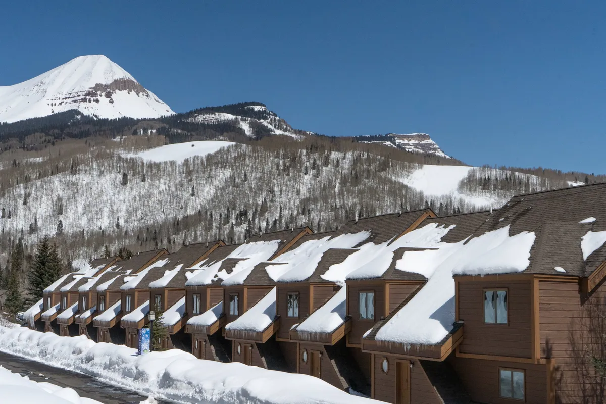 A picturesque row of snow-covered, brown townhome-style accommodations sits at the base of dramatic snowy mountain peaks and ski slopes. The clear blue sky overhead creates an inviting winter resort atmosphere, and balconies offer views of the slopes. The setting suggests immediate access to outdoor adventures and cozy retreat after activities.