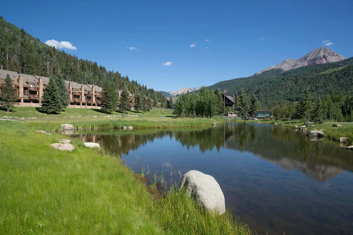A reflective pond bordered by natural rocks sits before a row of rustic mountain lodges, set against a backdrop of forested mountains and an expansive blue sky. The scene is peaceful and inviting, offering a sense of retreat and connection to the outdoors.