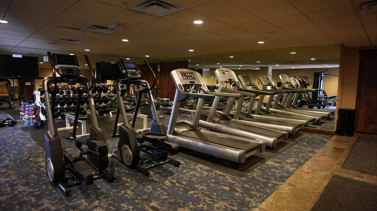 A bright, modern fitness center featuring a row of treadmills and elliptical machines facing large windows, with a well-organized dumbbell rack and exercise mats visible. The space is inviting and encourages guests to maintain their workout routines while traveling.