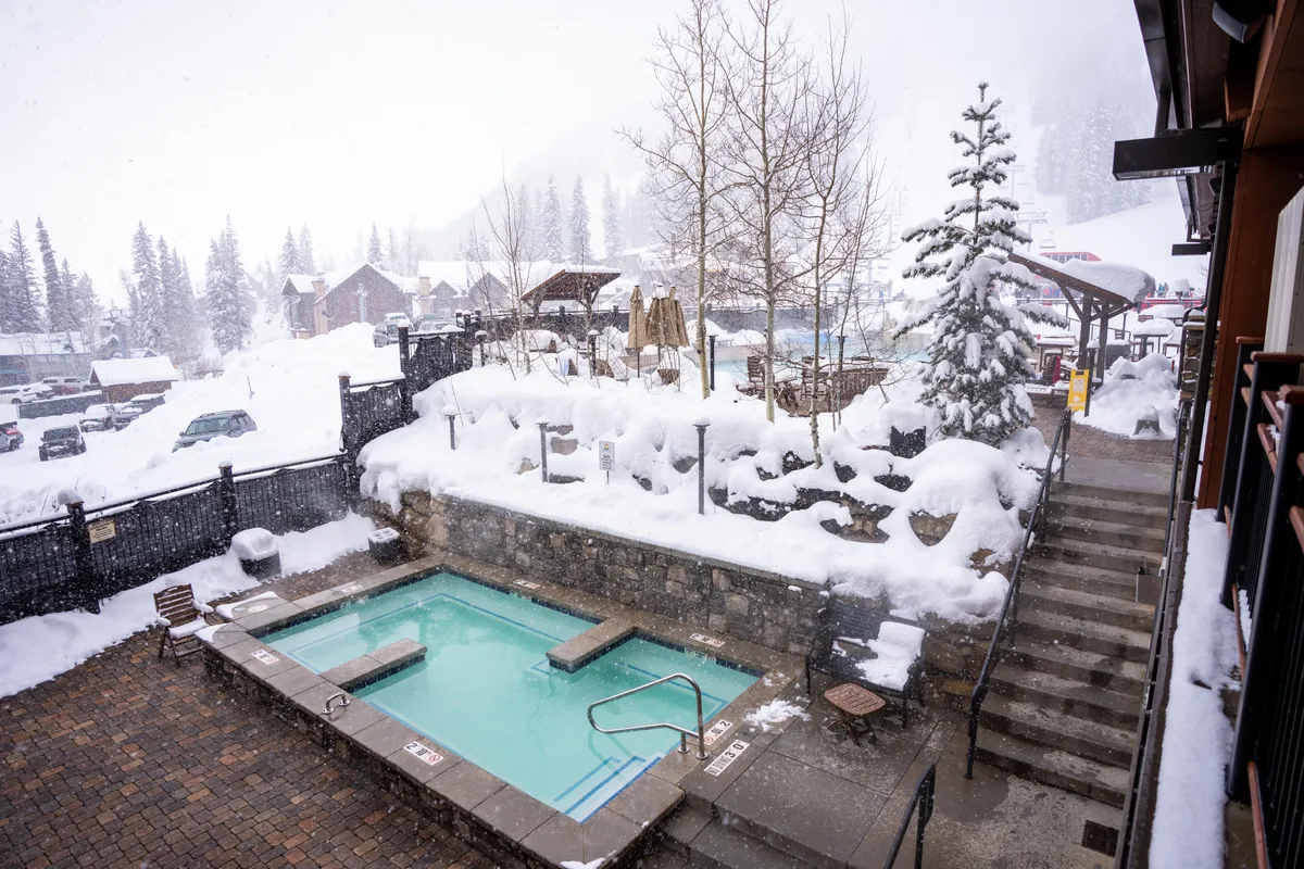 A heated outdoor pool with crystal-clear turquoise water set against a snowy landscape. Snow-laden trees and umbrellas surround the area, and stairs lead up to higher outdoor terraces. The setting is both luxurious and inviting, offering a warm retreat amid winter scenery.