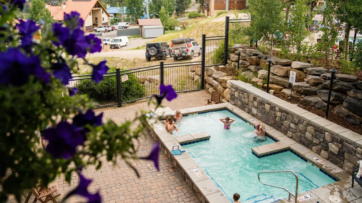 A heated outdoor pool surrounded by a stone wall, with people relaxing and enjoying the warm water. Lush purple flowers in the foreground add a burst of color, and the tranquil mountain backdrop offers a sense of relaxation and leisure after a day of adventure.