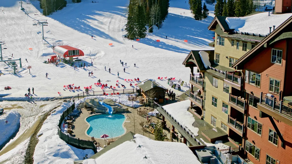 A lively winter resort scene featuring skiers on snowy slopes, a heated outdoor pool with lounge chairs, and a multi-story lodge with balconies. The ski lift and a red equipment building are visible, illustrating easy ski access and a festive resort environment.