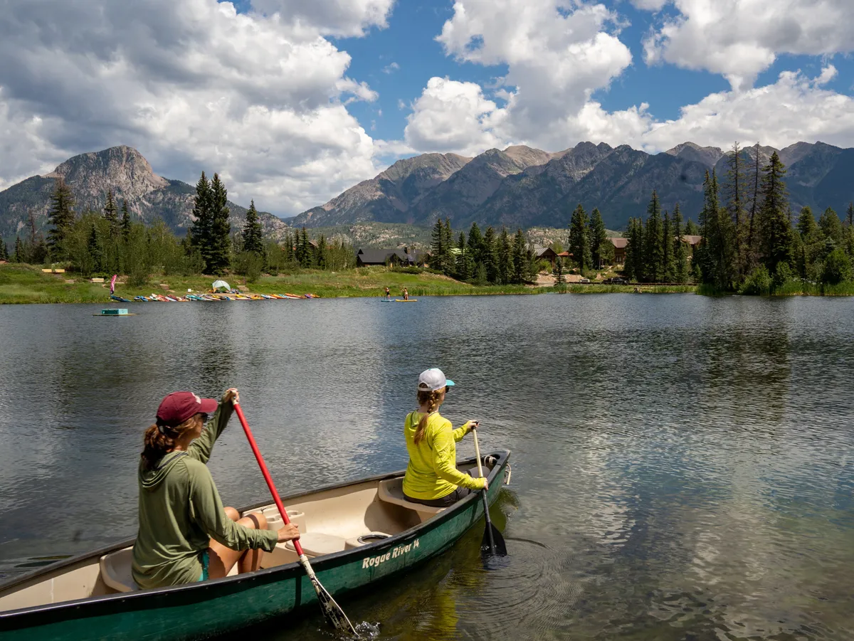 Two people paddle a canoe on a calm alpine lake, surrounded by a mountain range and forested lakeshore with hints of rustic buildings. The serene water reflects the sky and mountains, blending adventure with peaceful wilderness.