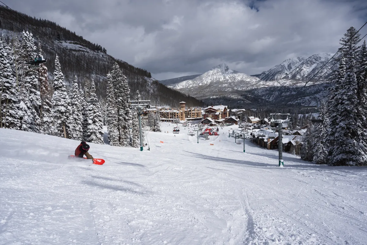 A snowboarder carves down a snowy slope surrounded by tall, snow-covered pine trees, with ski lift chairs and poles visible in the background. At the base, mountain resort buildings are nestled amidst the winter landscape, capturing the thrill and accessibility of mountain sports.